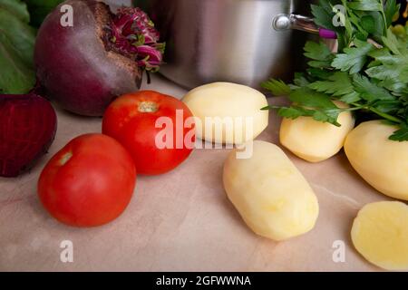 Rohzutaten zur Herstellung von Rotrüben-Borscht - frisches Gemüse, Rüben, Tomaten, Kartoffeln und Kräuter auf einem Schneidebrett in der Nähe einer Edelstahlwanne auf t Stockfoto