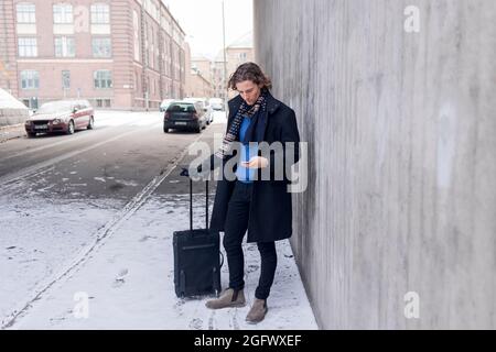 Junger Mann mit Koffer, der das Telefon auf dem verschneiten Bürgersteig überprüft Stockfoto