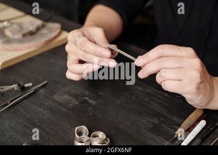 Nahaufnahme einer nicht erkennbaren Juwelierin, die am Schreibtisch sitzt und mit einer Handfeile einen handgefertigten Ring herstellt Stockfoto