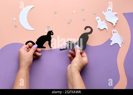 Niedliche Halloween-Hintergrund mit Copy-Space. Hände mit schwarzen Kawaii-Katzen und niedlichen weißen Papiergeistern. Flach auf rosa und lila Papier legen mit Stockfoto