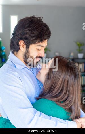 Vertikale Aufnahme eines fröhlichen jungen hispanischen Paares, das sich in Innenräumen umarmt und anschaut Stockfoto