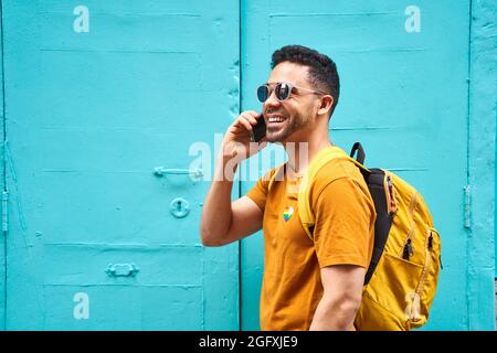 Ein fröhlicher junger Latino-Mann mit seinem Rucksack beantwortet sein Handy und geht die Straße entlang, trägt ein orangefarbenes T-Shirt mit lgtbi-Aufkleber und Sonnenbrille. Hochwertige Fotos Stockfoto