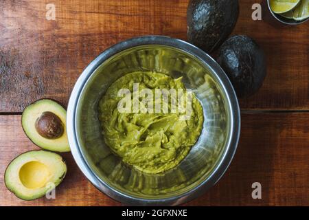 Draufsicht auf eine Metallschale mit frischer Guacamole darin und reifen Avocados daneben auf einem Holztisch Stockfoto