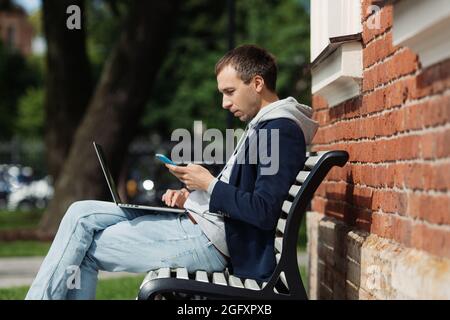 Millennial Geschäftsmann Multitasking lesen E-Mail im Smartphone verwenden Laptop sitzen auf der Bank im Freien Stockfoto