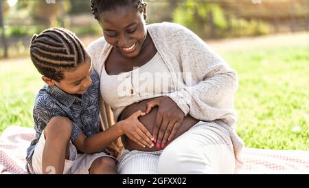 Glückliche afrikanische Familie erwartet anderes Baby - Afro Mutter und Sohn berühren den schwanger Bauch dabei Herzform mit den Händen Stockfoto