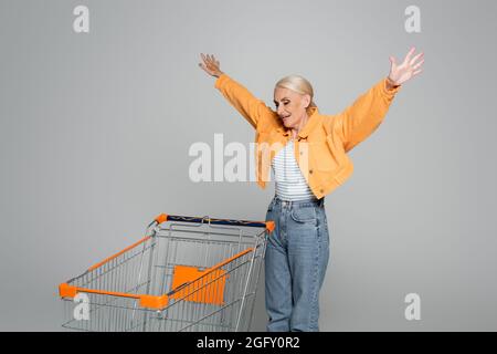 Aufgeregt ältere Frau zeigt Wow-Geste, während Blick auf den Warenkorb auf grau Stockfoto