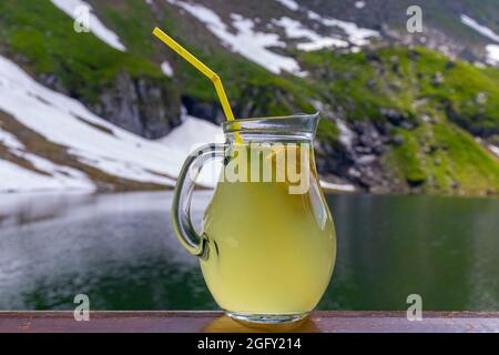 Ein Becher mit Limonade und Stroh auf einem Tisch mit verschwommener Sicht auf einen See in den Bergen im Hintergrund. Foto eines Glases mit Limonade. Stockfoto