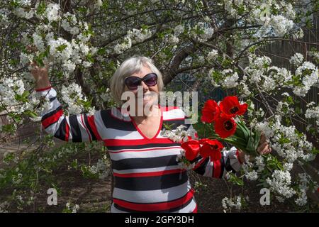 Halblanges Porträt einer lächelnden älteren Frau, die unter einem blühenden Kirschbaum mit roten Tulpen in ihren Händen steht. Stockfoto