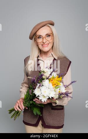 Stilvolle ältere Frau lächelt, während sie Blumen auf Grau isoliert hält Stockfoto