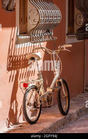 Faltfahrrad, das einen Schatten außerhalb eines griechischen Hauses wirft, typisch griechisch, typisch griechisch, Fahrrad am Fenster lehnend, Fahrrad an einer Wand lehnend. Stockfoto