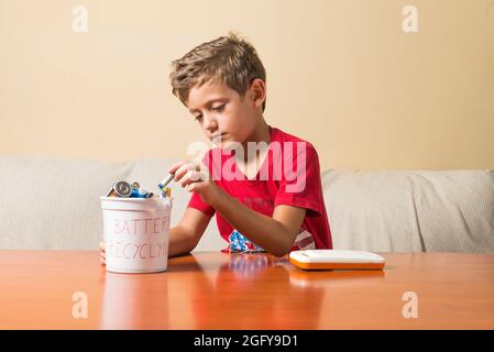 Ein Kind, das einen leeren Akku aus einem Spielzeug herausgenommen und in einen Recyclingbehälter gestellt hat. Konzept der Erziehung von Kindern in Umweltwerten. Stockfoto