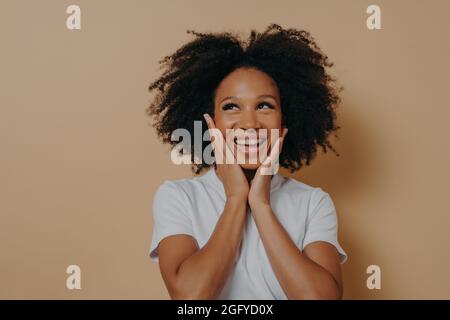 Fröhliche junge dunkelhäutige Frau, die sich glücklich fühlt, lächelt der Kamera zu und berührt Wangen mit beiden Händen Stockfoto