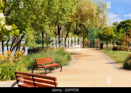 Parkovye Holzbänke auf dem gepflasterten Bürgersteig des Dnipo-Damms vor dem Hintergrund einer Sommerlandschaft und eines städtischen grünen Parks in Kiew. Stockfoto