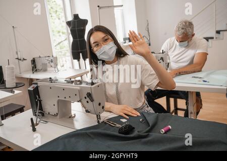 Asiatische Näherin in medizinische Maske winkende Hand in der Nähe Nähmaschine und Kollegen Stockfoto