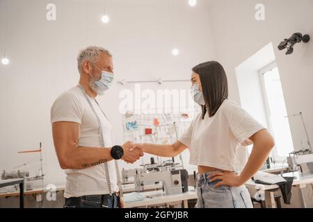 Seitenansicht von multiethnischen Designern in medizinischen Masken, die sich im Werk die Hände schütteln Stockfoto