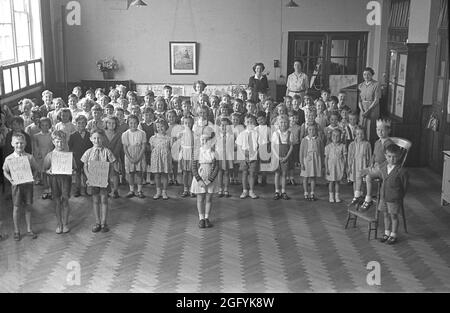 1955, historische, kleine Kinder in einer Versammlung an einer Grundschule, England, Großbritannien. Das Bild zeigt Schülerinnen und Schüler, die als Gruppe zusammenstehen, mit drei Jungen an der Vorderseite, die Karten hochhalten und sagen: „Tailor Coat 1D“ und „Hatter hat 2D“, „Cobbler Shoes 3D“, vielleicht als Teil einer Aufführung oder eines Spiels, Wie ein Junge sitzt auf einem großen Stuhl mit einer Krone auf dem Kopf, der König? Stockfoto