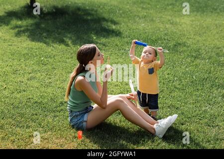 Junge Mutter sitzt auf Gras und bläst Seifenblasen in der Nähe Kleinkind Sohn Stockfoto