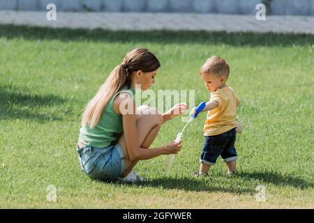 Junge Mutter hält Seifenblasenflasche in der Nähe Kleinkind Sohn auf Gras Stockfoto