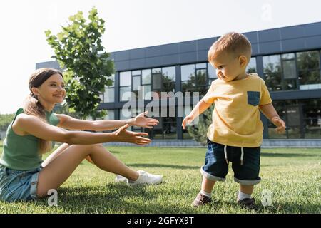 Fröhliche Mutter mit ausgestreckten Händen, die auf Gras sitzt und den Kleinkind draußen erreicht Stockfoto