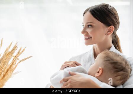 Lächelnde Mutter umarmt Kleinkind Sohn mit geschlossenen Augen im Bademantel Stockfoto