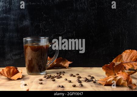 Ein Glas aus hartgebrühtem Kaffee mit Kaffeebohnen und Herbstblättern auf einem Holztisch. Selektiver Fokus. Stockfoto