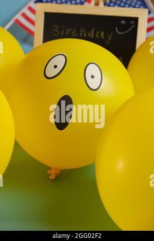 Gelbe Emoji-Ballons auf dem Hintergrund der Tafel mit der Ereignisbeschriftung und amerikanischen Flaggen. Geburtstag des Emoticons. Stockfoto
