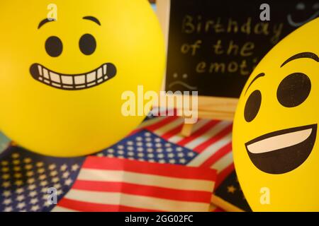Gelbe Emoji-Ballons auf dem Hintergrund der Tafel mit der Ereignisbeschriftung und amerikanischen Flaggen. Geburtstag des Emoticons. Stockfoto