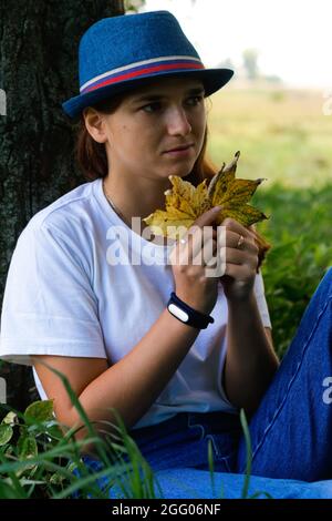 Unschärfe-Porträt einer traurigen, verträumten jungen Frau mit braunen Haaren, die einen Hut im Freien trägt. Weibliche Hand mit gelben trockenen Blättern. Hallo Herbst. Frauen sind in Seatin Stockfoto