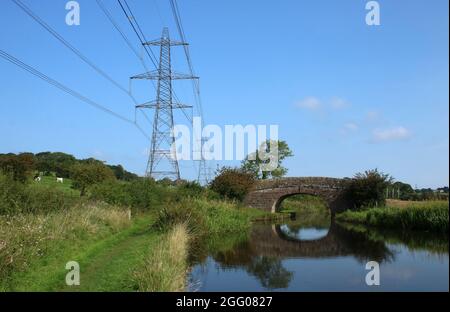Lancaster-Kanal, steinerne Bogenbrücke mit Reflexionen in Wasserstraße und Pylon und Stromleitungen gegen blauen Himmel gesehen. Stockfoto