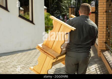 Der Typ trägt zwei Holzbänke. Der Mann zieht Objekte. Vorbereitung der Veranstaltung. Ein Mitarbeiter schleppt im Sommer Möbel. Stockfoto