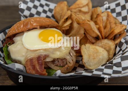 Gebratenes Ei auf diesem beladenen Speck- und Käseburger, serviert mit Kartoffelbüffelchips für eine appetitliche Mahlzeit. Stockfoto