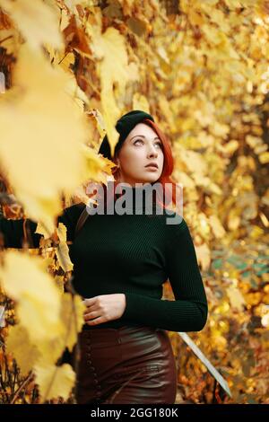 Schöne junge rothaarige Frau im Herbstpark. Stilvolles Modell zwischen gelben, trockenen Laub von Bäumen. Tolle Frau in schwarzer Baskenmütze, gestrickter Pullover schaut auf den Himmel. Spaziergang in der herbst Natur. Stockfoto