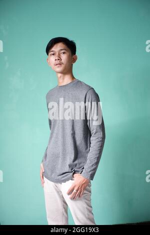 Portrait eines hübschen asiatischen Mannes mit grauem T-Shirt in cooler Pose. Dünner Mann auf grünem Hintergrund. Mann, der seine Hände mit einem s in seine Hosentasche legt Stockfoto