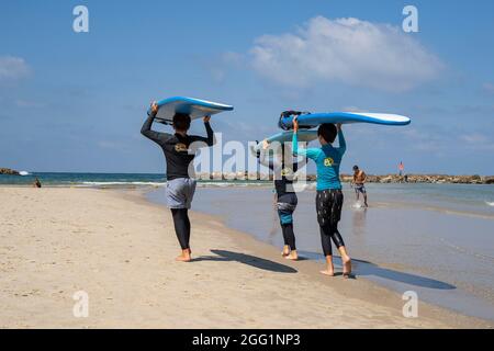 Tel Aviv, Israel - 15. August 2021: Drei junge Surfer, die ihre Bretter über den Kopf halten, fahren in Tel Aviv ins Meer. Stockfoto