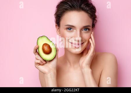 Lächelnde junge Frau mit einer halben Avocado. Foto von attraktiven Frau mit perfektem Make-up auf rosa Hintergrund. Beauty & Hautpflege Konzept Stockfoto