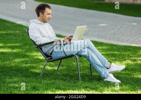 Blogger Mann arbeitet am Laptop im öffentlichen Park, verarbeitet Videos für Social-Media-Inhalte, sitzt im Stuhl Stockfoto