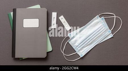 School Covid 19 Schutzkonzept. Schnelltest, Notizbücher und medizinische Schutzmaske auf grauem Hintergrund, Draufsicht, Zurück zur Schule, Coronav Stockfoto