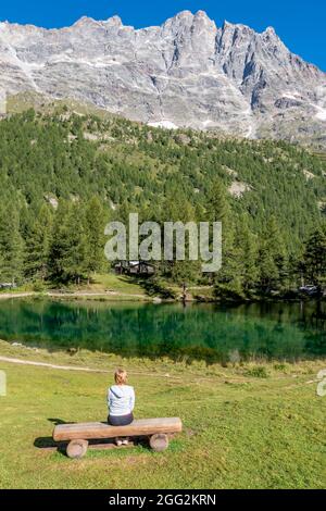 Eine blonde Frau, die auf einer Holzbank sitzt, bewundert das Panorama des Blauen Sees, in dem sich die umliegenden Berge spiegeln, das Aostatal, Italien Stockfoto