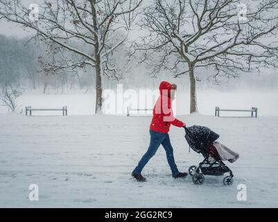 Verschneite Winterszene mit einer Mutter, die einen Kinderwagen schiebt, während sie an einem verschneiten Wintermorgen im Park spazieren geht. Straßenfotografie Saisonale Szene mit Schneefall in Stockfoto