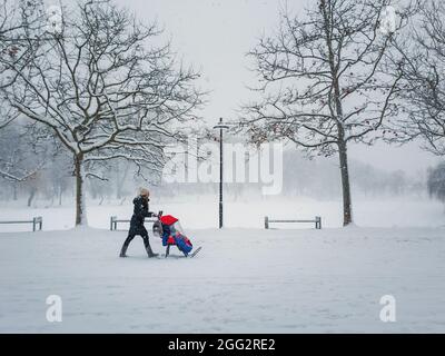 Verschneite Winterszene mit einer Mutter, die mit ihrem Baby auf einem Kinderwagen im Park spazieren geht. Straßenfotografie Saisonale Szene, flauschige Flocken Schneefall in der Stockfoto