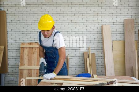 Der asiatische Zimmermann trägt einen gelben Harthut und schneidet in der Holzbearbeitungsanlage mit einer elektrischen Handsäge Sperrholz. Ein Schreibtisch mit Handwerkzeugen und Holzpfählen. Stockfoto