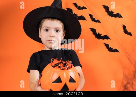 Halloween Kind. Ein Vorschuljunge in einem Hexenkostüm hält einen Kürbis mit Süßigkeiten auf einem orangen Hintergrund. Rabatte und Verkäufe für den Urlaub Stockfoto