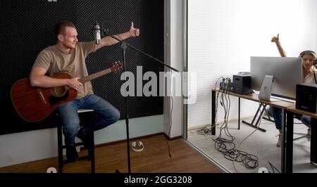 Der junge kaukasische Künstler mit Akustikgitarre überprüft die Korrektheit des Audiorecorders, bevor er mit dem Toningenieur beginnt. Musiker produzieren Stockfoto