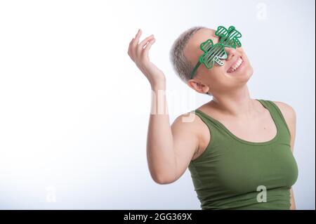 Porträt einer jungen kaukasischen Frau mit einem Männerhaarschnitt in einem grünen T-Shirt und einer fröhlichen Brille auf weißem Hintergrund. Das Mädchen feiert St. patrick Stockfoto