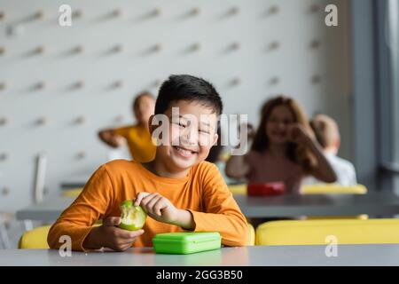 Glückliches asiatisches Kind, das mit frischem Apfel lächelt, vor der Kamera bei verschwommenen Kindern in der Schulkantine Stockfoto