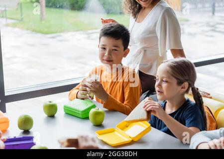 Glücklicher asiatischer Junge hält Sandwich während des Mittagessens in der Nähe eines Schulmädchen und einer afroamerikanischen Lehrerin Stockfoto