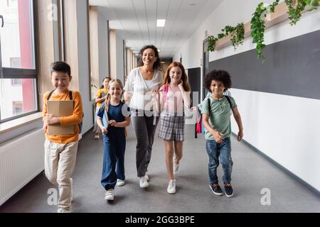 Glücklicher afroamerikanischer Lehrer, der im Schulkorridor mit multiethnischen Schülern läuft Stockfoto