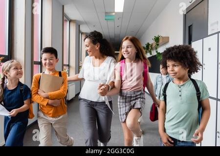 Fröhliche multikulturelle Schüler, die mit afroamerikanischem Lehrer entlang des Schulkorridors gehen Stockfoto