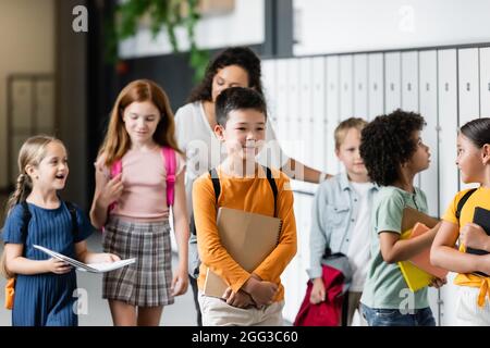 Multiethnische Schüler, die entlang des Schulkorridors in der Nähe einer verschwommenen afroamerikanischen Lehrerin gehen Stockfoto