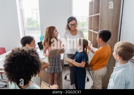 Multiethnische Schüler mit Büchern, die im Klassenzimmer mit einem glücklichen afroamerikanischen Lehrer sprechen Stockfoto